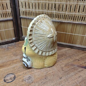 May include: A ceramic Tanuki figurine, a traditional Japanese raccoon dog, with a large hat and a sake bottle. The figurine is tan and cream-colored, with detailed carving and a whimsical design. The figurine is on a wooden surface.