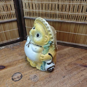 May include: A ceramic Tanuki statue, a traditional Japanese raccoon dog figure. The statue is yellow, white, and green, with a large belly, a hat, and a sake bottle. It is a decorative item.
