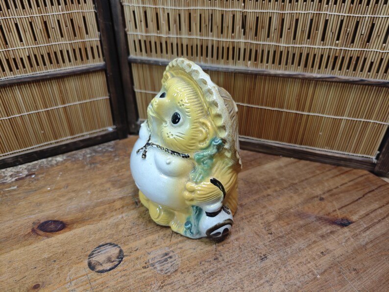 May include: A ceramic Tanuki statue, a traditional Japanese raccoon dog figure. The statue is yellow, white, and green, with a large belly, a hat, and a sake bottle. It is a decorative item.
