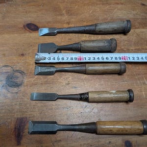 Puede incluir: Cinco cinceles de madera antiguos con hojas de metal, dispuestos sobre una superficie de madera. Una cinta m&eacute;trica se coloca sobre los cinceles. Los cinceles tienen mangos de madera y hojas de metal de varias formas y tama&ntilde;os.
