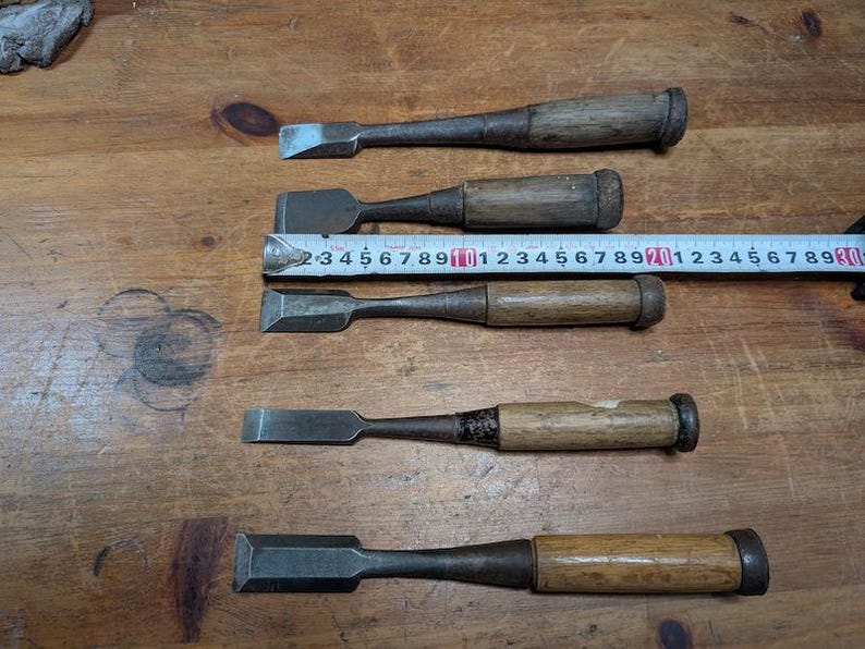 Puede incluir: Cinco cinceles de madera antiguos con hojas de metal, dispuestos sobre una superficie de madera. Una cinta m&eacute;trica se coloca sobre los cinceles. Los cinceles tienen mangos de madera y hojas de metal de varias formas y tama&ntilde;os.