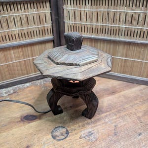 May include: A wooden Japanese garden lantern with a hexagonal top and a square finial. The lantern has a dark brown finish and is set on a wooden surface. The background features a bamboo screen.