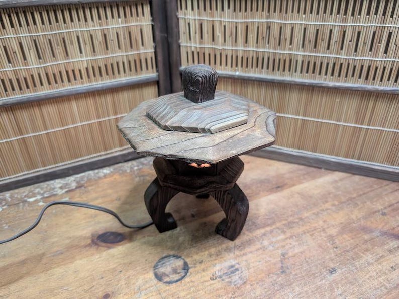 May include: A wooden Japanese garden lantern with a hexagonal top and a square finial. The lantern has a dark brown finish and is set on a wooden surface. The background features a bamboo screen.