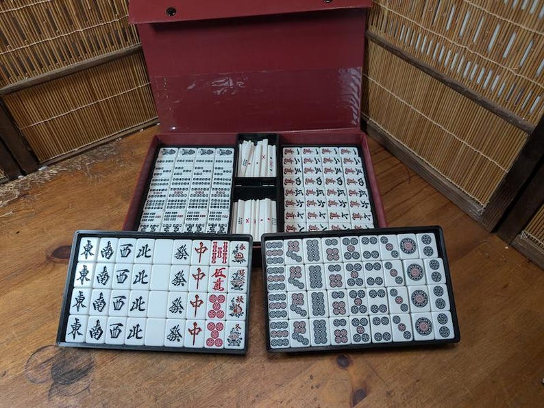Op de afbeelding: Een complete Mahjong-set met witte tegels met zwarte en rode symbolen, in zwarte trays en een rode koffer. De set is open en toont de tegels en de koffer.