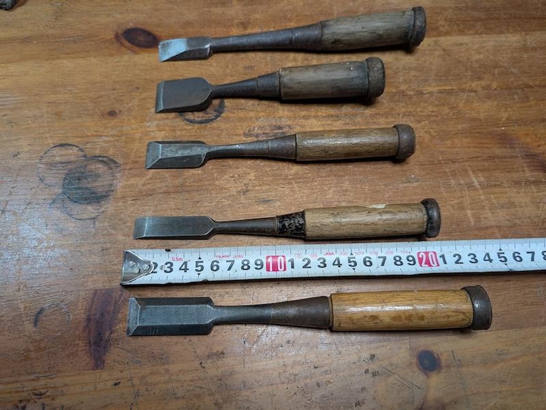 Puede incluir: Cinco cinceles de madera antiguos con hojas de metal, dispuestos sobre una superficie de madera. Una cinta m&eacute;trica se coloca junto a los cinceles. Los cinceles tienen mangos de madera y hojas de metal de varias formas y tama&ntilde;os.