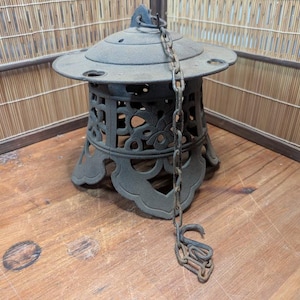May include: An antique, weathered, cast iron Japanese garden lantern. The lantern has a round top with holes, a decorative openwork body, and a chain. The lantern is dark gray and sits on a wooden surface.