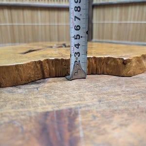 May include: A natural wood slab with a live edge, approximately 3 inches thick, is measured with a metal ruler. The wood has a light brown color with darker grain patterns and a unique, organic shape. The slab is likely for decorative or functional use.
