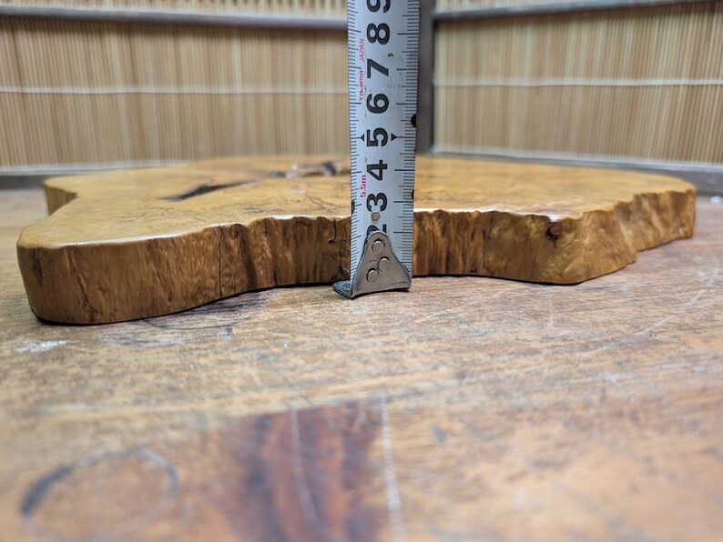 May include: A natural wood slab with a live edge, approximately 3 inches thick, is measured with a metal ruler. The wood has a light brown color with darker grain patterns and a unique, organic shape. The slab is likely for decorative or functional use.