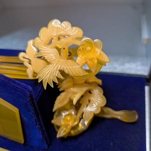 May include: A detailed, pale yellow hair comb with a bird and floral design. The comb is carved with intricate details, featuring a bird in flight and delicate flowers. The comb is set against a blue velvet background, suggesting a vintage or antique piece.