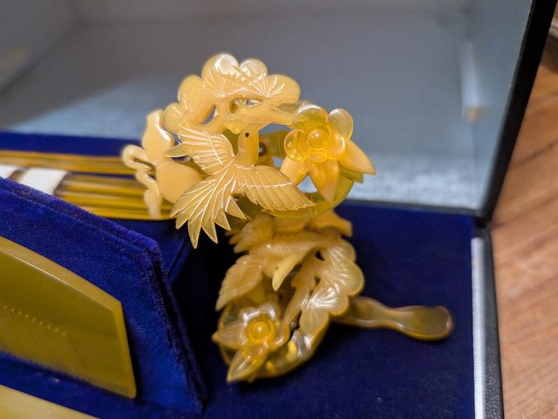 May include: A detailed, pale yellow hair comb with a bird and floral design. The comb is carved with intricate details, featuring a bird in flight and delicate flowers. The comb is set against a blue velvet background, suggesting a vintage or antique piece.