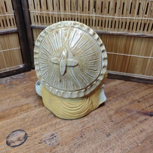 May include: A ceramic figurine of a seated figure with a large, round, sun-like hat. The hat is beige with carved radial lines and a cross-shaped detail. The figure has yellow hair and a white face. The figurine is on a wooden surface.