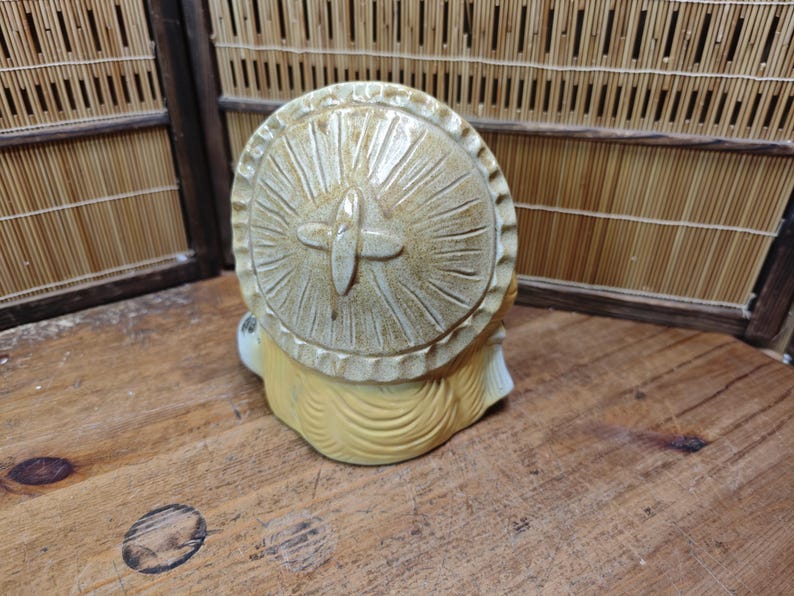 May include: A ceramic figurine of a seated figure with a large, round, sun-like hat. The hat is beige with carved radial lines and a cross-shaped detail. The figure has yellow hair and a white face. The figurine is on a wooden surface.