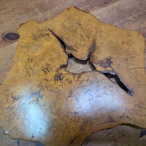 May include: A uniquely shaped, natural wood slab with a light brown color and a prominent knot. The slab has a rough, organic edge and a large, irregular opening in the center. The wood grain is visible throughout the surface.