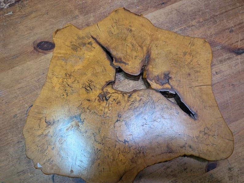 May include: A uniquely shaped, natural wood slab with a light brown color and a prominent knot. The slab has a rough, organic edge and a large, irregular opening in the center. The wood grain is visible throughout the surface.