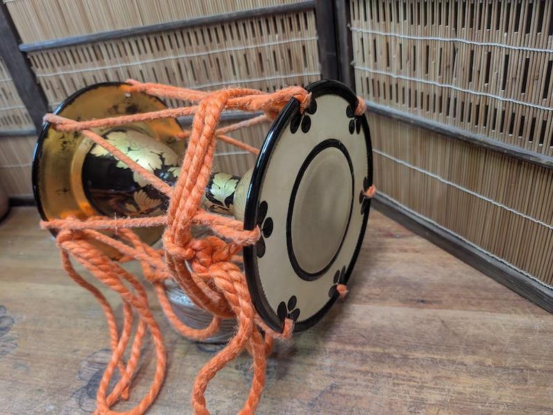 K&ouml;nnte beinhalten: Traditionelle japanische Trommel mit zylindrischem Korpus und dekorativen goldenen und schwarzen Akzenten. Die Trommelfelle sind beige mit schwarzen floralen Designs. Helles orangefarbenes Seil sichert die Trommelstruktur.