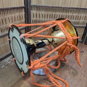 K&ouml;nnte beinhalten: Eine japanische Tsuzumi-Trommel mit einem schwarzen und goldenen Korpus, verziert mit floralen Mustern. Die Trommelfelle sind cremefarben mit schwarzen Akzenten, und die Trommel ist mit orangefarbenem Seil bespannt. Die Trommel liegt auf einer Holzoberfl&auml;che.