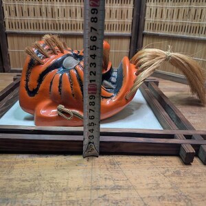 May include: An orange and black Japanese Noh mask with a fierce expression, featuring a long, flowing hairpiece. The mask is displayed on a wooden frame, with a ruler next to it for scale. The mask is approximately 6 inches tall.