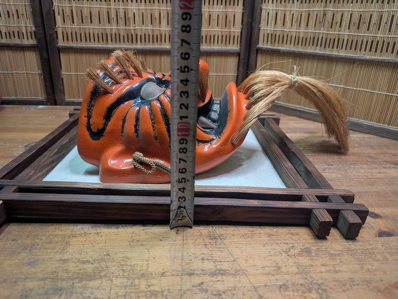 May include: An orange and black Japanese Noh mask with a fierce expression, featuring a long, flowing hairpiece. The mask is displayed on a wooden frame, with a ruler next to it for scale. The mask is approximately 6 inches tall.