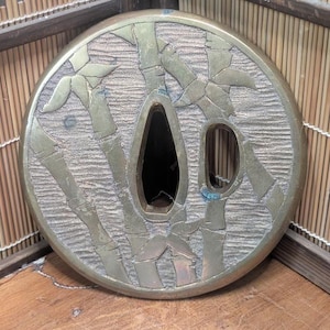 May include: A round, antique metal tsuba, a Japanese sword guard, with a bamboo design. The tsuba has a central opening and two smaller openings. The bamboo stalks and leaves are raised against a textured background. The metal has a tarnished, aged appearance.