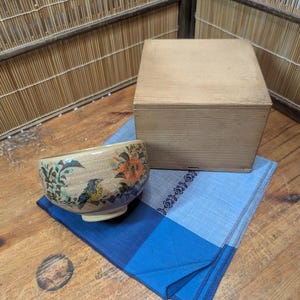 Puede incluir: Un cuenco de cerámica beige con diseños florales y de pájaros, una caja de madera y un paño con estampado azul y blanco están dispuestos sobre una superficie de madera. El cuenco presenta detalles en naranja y azul.