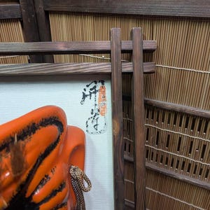 May include: An orange and black ceramic mask with a rope detail, displayed in front of a framed artwork with Japanese calligraphy. The background features a bamboo screen and wooden supports, creating a traditional aesthetic.