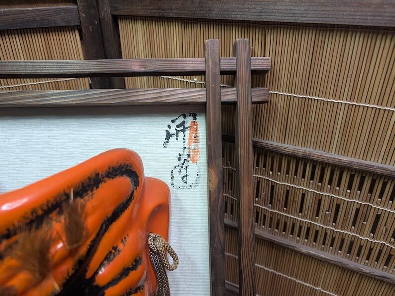 May include: An orange and black ceramic mask with a rope detail, displayed in front of a framed artwork with Japanese calligraphy. The background features a bamboo screen and wooden supports, creating a traditional aesthetic.