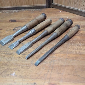 Może przedstawiać: Pięć zabytkowych dłut z drewnianymi uchwytami ułożonych na drewnianej powierzchni. Dłuta mają metalowe ostrza i drewniane uchwyty. Ostrza są srebrne, a uchwyty jasnobrązowe. Narzędzia te są prawdopodobnie używane do obróbki drewna.