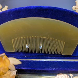May include: A light amber comb with fine teeth, set against a deep blue velvet backdrop. The comb has a curved top and two small, decorative metal accents. The design suggests a focus on hair care and grooming.