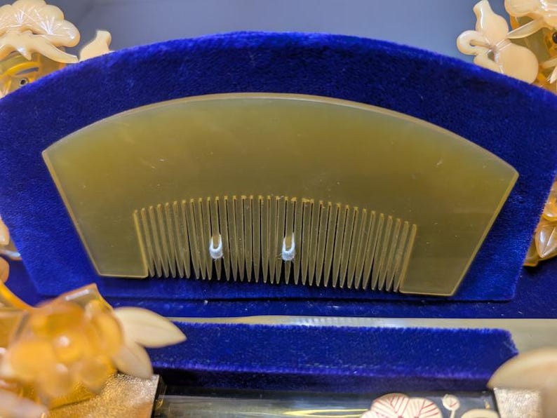 May include: A light amber comb with fine teeth, set against a deep blue velvet backdrop. The comb has a curved top and two small, decorative metal accents. The design suggests a focus on hair care and grooming.
