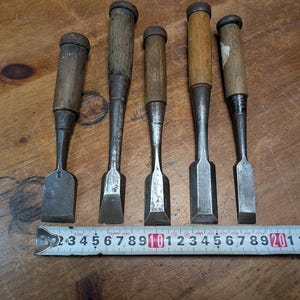 Puede incluir: Cinco cinceles de madera antiguos con hojas de metal y mangos de madera est&aacute;n dispuestos sobre una superficie de madera. Una cinta m&eacute;trica se coloca debajo. Los cinceles var&iacute;an en tama&ntilde;o y forma, mostrando signos de desgaste.