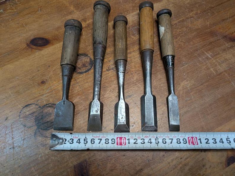 Puede incluir: Cinco cinceles de madera antiguos con hojas de metal y mangos de madera est&aacute;n dispuestos sobre una superficie de madera. Una cinta m&eacute;trica se coloca debajo. Los cinceles var&iacute;an en tama&ntilde;o y forma, mostrando signos de desgaste.