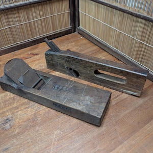 Op de afbeelding: Twee antieke houten handschaven, waarschijnlijk voor houtbewerking. De schaven zijn donkerbruin met zichtbare slijtage, wat duidt op leeftijd en gebruik. Eén schaaf heeft een handvat en een mes, de andere heeft een platte bovenkant. De achtergrond omvat een houten oppervlak en een bamboe scherm.