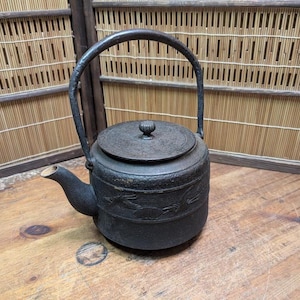 Puede incluir: Una tetera de hierro fundido negro con un asa curva y una tapa con un pequeño pomo. La tetera tiene un pico y está decorada con un diseño en relieve. La tetera está sobre una superficie de madera.