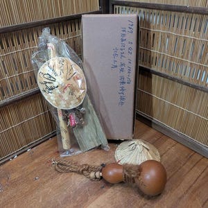 Puede incluir: Una colección de artesanías japonesas, que incluye un objeto en forma de calabaza con una cuerda, un pequeño sombrero tejido y un artículo empaquetado con un sombrero de bambú. También hay una caja de cartón con texto japonés.