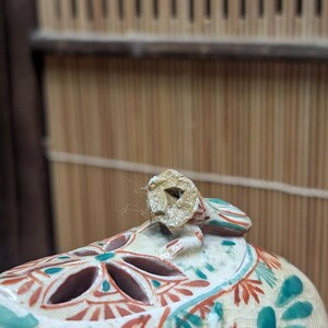 May include: Close-up of a ceramic decorative object with a lid. The lid has a small, broken handle. The object is painted with red, teal, and white floral patterns. The background is a bamboo screen.