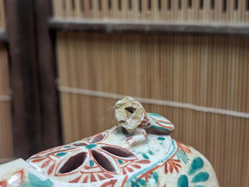 May include: Close-up of a ceramic decorative object with a lid. The lid has a small, broken handle. The object is painted with red, teal, and white floral patterns. The background is a bamboo screen.