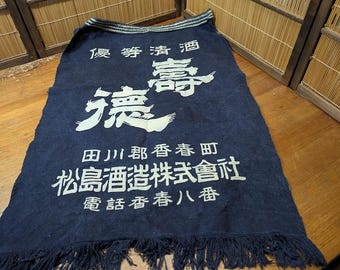 May include: A navy blue cloth with white Japanese characters and fringe along the bottom. The text includes Japanese characters and the words "Superior Sake" and "Matsushima Brewery Co., Ltd."