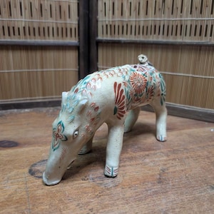 May include: A ceramic figurine of a boar, painted with floral patterns in red, green, and blue. The boar is cream-colored and stands on four legs, with its snout lowered. The figurine has a small tail and is set against a wooden surface.