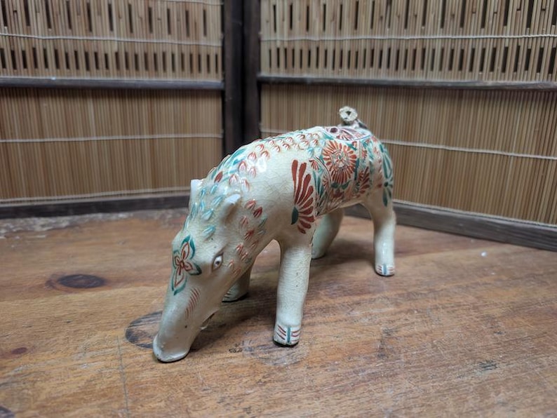 May include: A ceramic figurine of a boar, painted with floral patterns in red, green, and blue. The boar is cream-colored and stands on four legs, with its snout lowered. The figurine has a small tail and is set against a wooden surface.