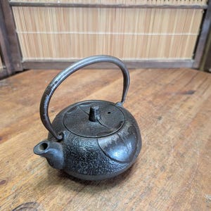 Könnte beinhalten: Kleine, schwarze Gusseisen-Teekanne mit gebogenem Griff und Deckel. Die Teekanne hat eine strukturierte Oberfläche und einen Ausguss. Der Hintergrund ist eine Holzoberfläche und ein Bambus-Sichtschutz.