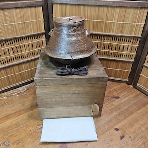 Puede incluir: Una olla de cerámica marrón rústica con tapa, apoyada sobre una caja de madera desgastada. Un cordón oscuro está atado en forma de lazo frente a la olla. El fondo presenta una pantalla de bambú y un suelo de madera.
