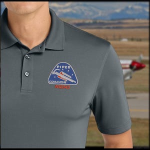 May include: A charcoal gray short-sleeve polo shirt featuring a triangular embroidered patch on the chest. The patch displays the text PIPER COMANCHE with a graphic of an airplane and stars. Below the patch, the text N12345 is embroidered in red.