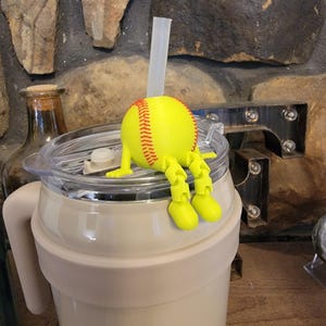 Può includere: Un bicchiere color crema con coperchio trasparente e un topper per cannuccia a tema baseball giallo. Il topper ha un design a palla da baseball con cuciture rosse e gambe e piedi gialli. Una cannuccia trasparente si estende dalla palla da baseball.