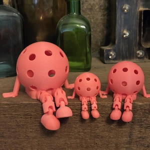 May include: Three coral-colored, spherical figures with multiple circular cutouts, articulated arms, and jointed legs. The figures are arranged in a row, with one large figure on the left, a small figure in the center, and a medium figure on the right.