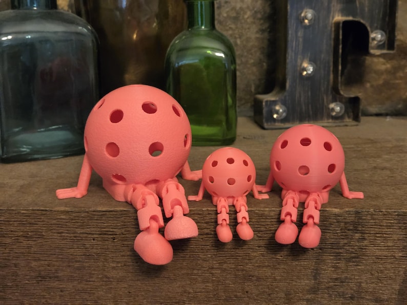 May include: Three coral-colored, spherical figures with multiple circular cutouts, articulated arms, and jointed legs. The figures are arranged in a row, with one large figure on the left, a small figure in the center, and a medium figure on the right.