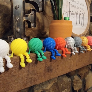 Op de afbeelding: Een rij kleurrijke golfbalfiguren met aangehechte benen, in wit, geel, groen, blauw, oranje, zilver, grijs en roze. De golfballen zitten op een houten oppervlak. De achtergrond omvat een decoratieve metalen letter en een bord.