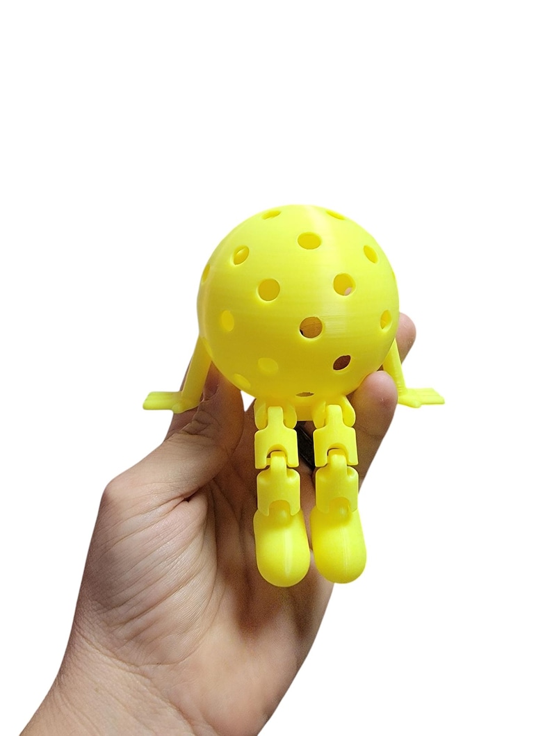 May include: A yellow 3D-printed pickleball with articulated legs and arms. The ball has several circular holes. The legs and arms are connected with hinges, allowing for movement. The object is held in a hand against a white background.