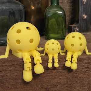 May include: Three bright yellow, spherical figures with multiple circular cutouts, articulated arms, and jointed legs. Each figure features a round head with a hollow, perforated design and dangling limbs, arranged in a row from largest to smallest.