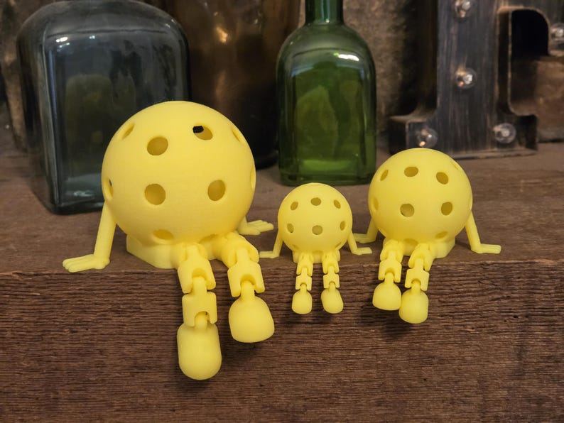 May include: Three bright yellow, spherical figures with multiple circular cutouts, articulated arms, and jointed legs. Each figure features a round head with a hollow, perforated design and dangling limbs, arranged in a row from largest to smallest.