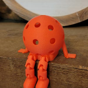 May include: An orange, spherical, 3D-printed figure with a perforated surface, articulated legs, and small arms. The figure is sitting on a wooden surface. The design resembles a whimsical character or toy.